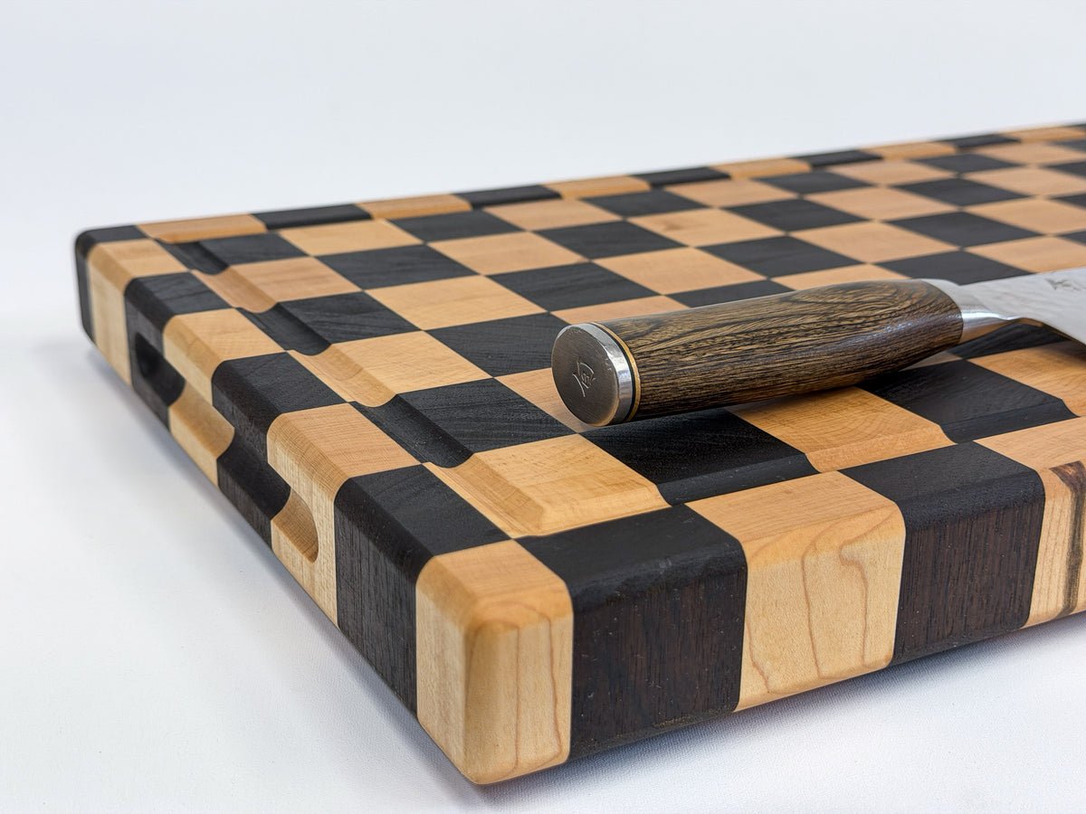 Close-up of the maple grain, dark checkerboard pattern, rounded corner and side grip of a handcrafted cutting board.