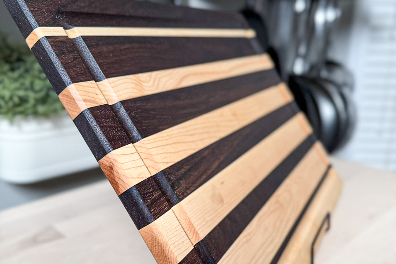 A striped wooden cutting board with alternating light and dark wood bands, resting on a kitchen counter with hanging pots and pans in the background. zoomed in...close up grain shot