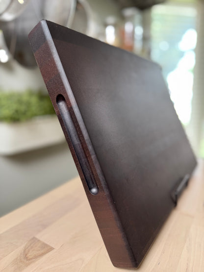 Dark brown end grain walnut cutting board on a wooden surface with a blurred kitchen background