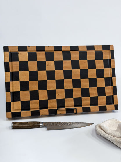 Handcrafted maple checkerboard cutting board displayed upright with a chef’s knife and folded kitchen towel.