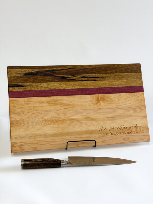 Portrait view of a handcrafted maple, purpleheart and black limba charcuterie board displayed with a chef’s knife.