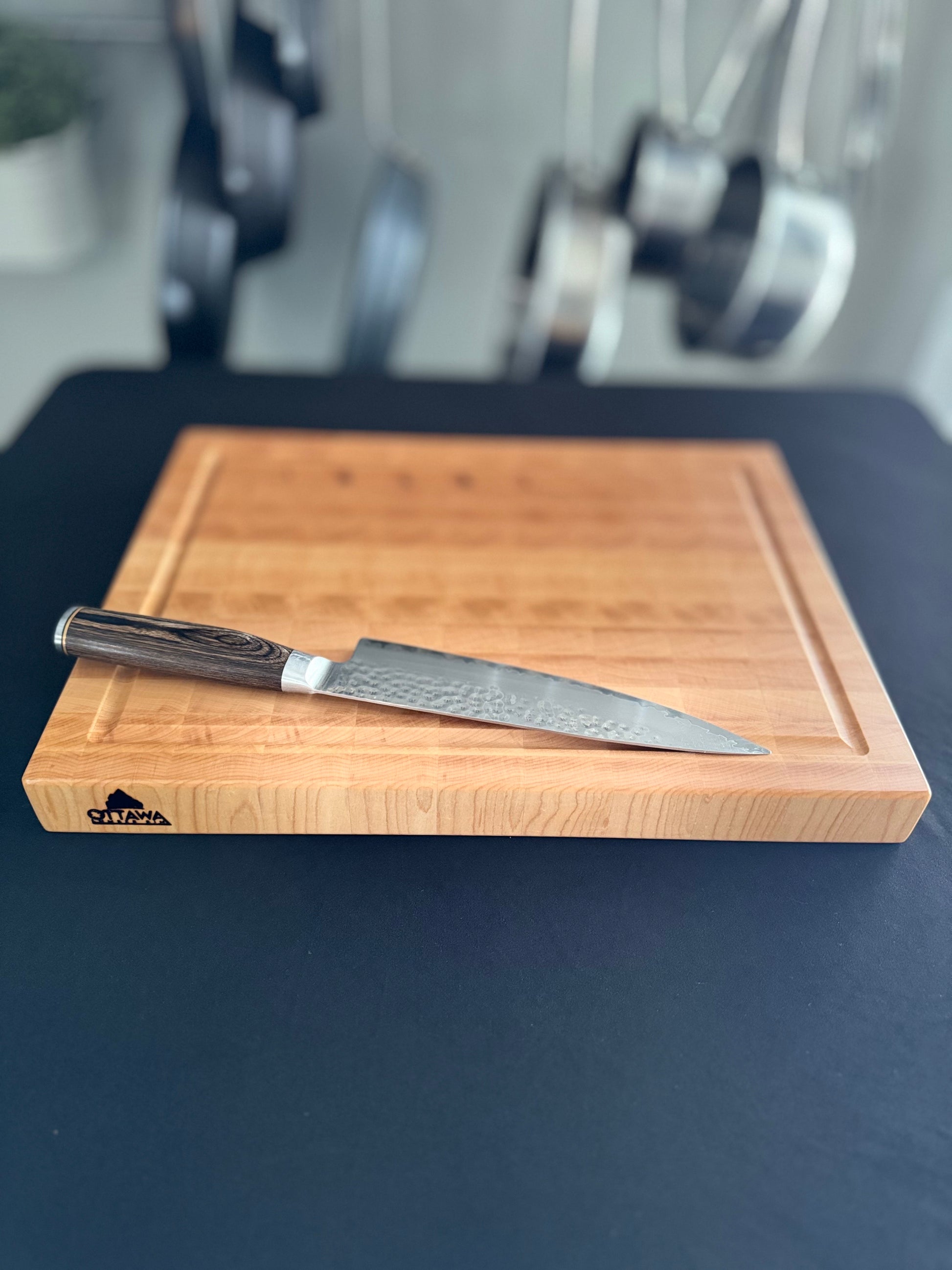 An end grain maple cutting board laying flat on a black tabletop with a knife on the board for scale.