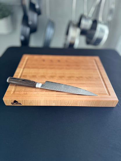 An end grain maple cutting board laying flat on a black tabletop with a knife on the board for scale.