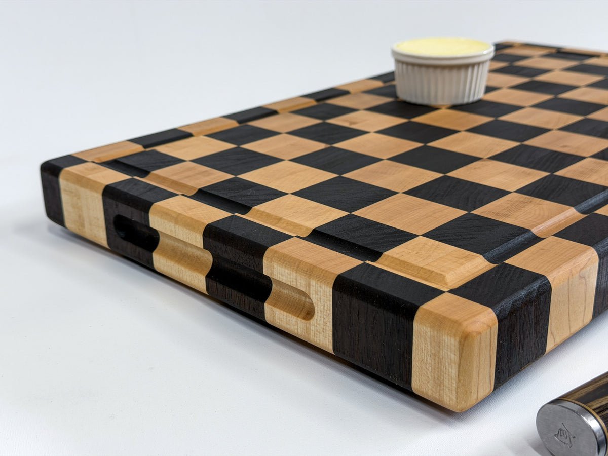 Close-up of a thick maple checkerboard cutting board with a juice groove, side grips and ramekin of butter.