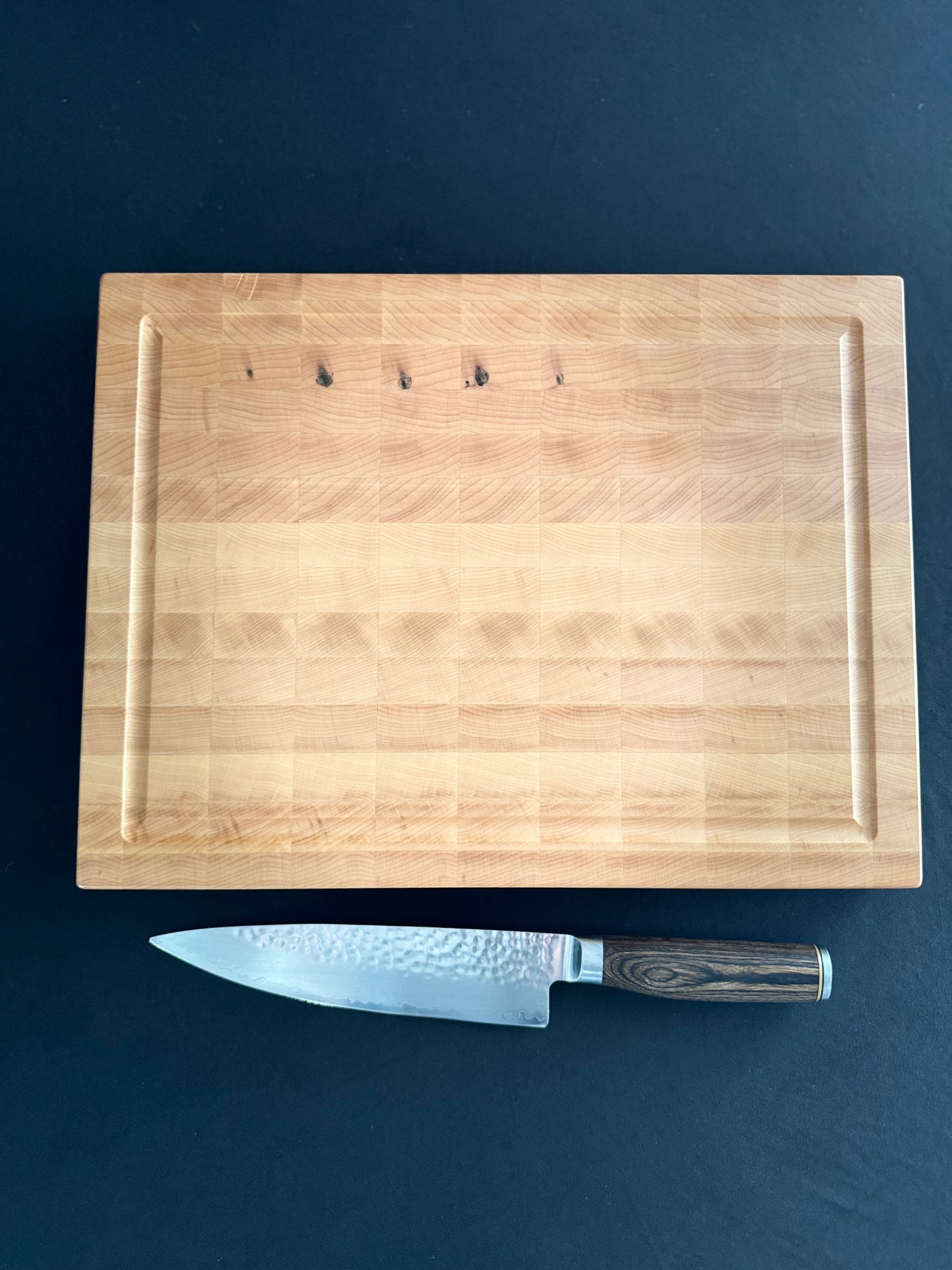 An end grain maple cutting board laying flat on a black tabletop with a knife besidethe board for scale.
