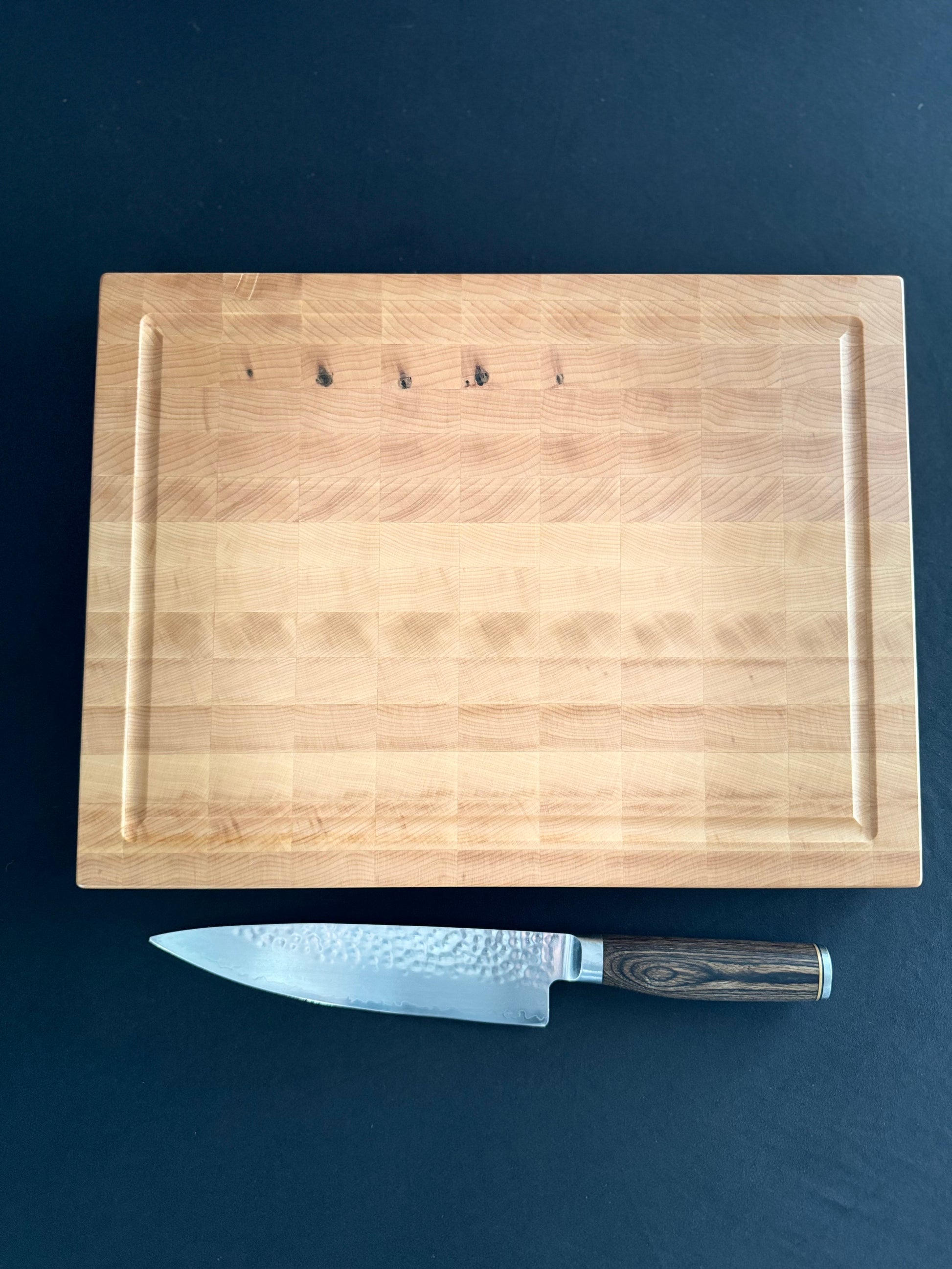 An end grain maple cutting board laying flat on a black tabletop with a knife besidethe board for scale.