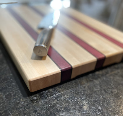 Maple edge grain cutting board with Purpleheart and walnut accent stripes displayed with a chef's knife.