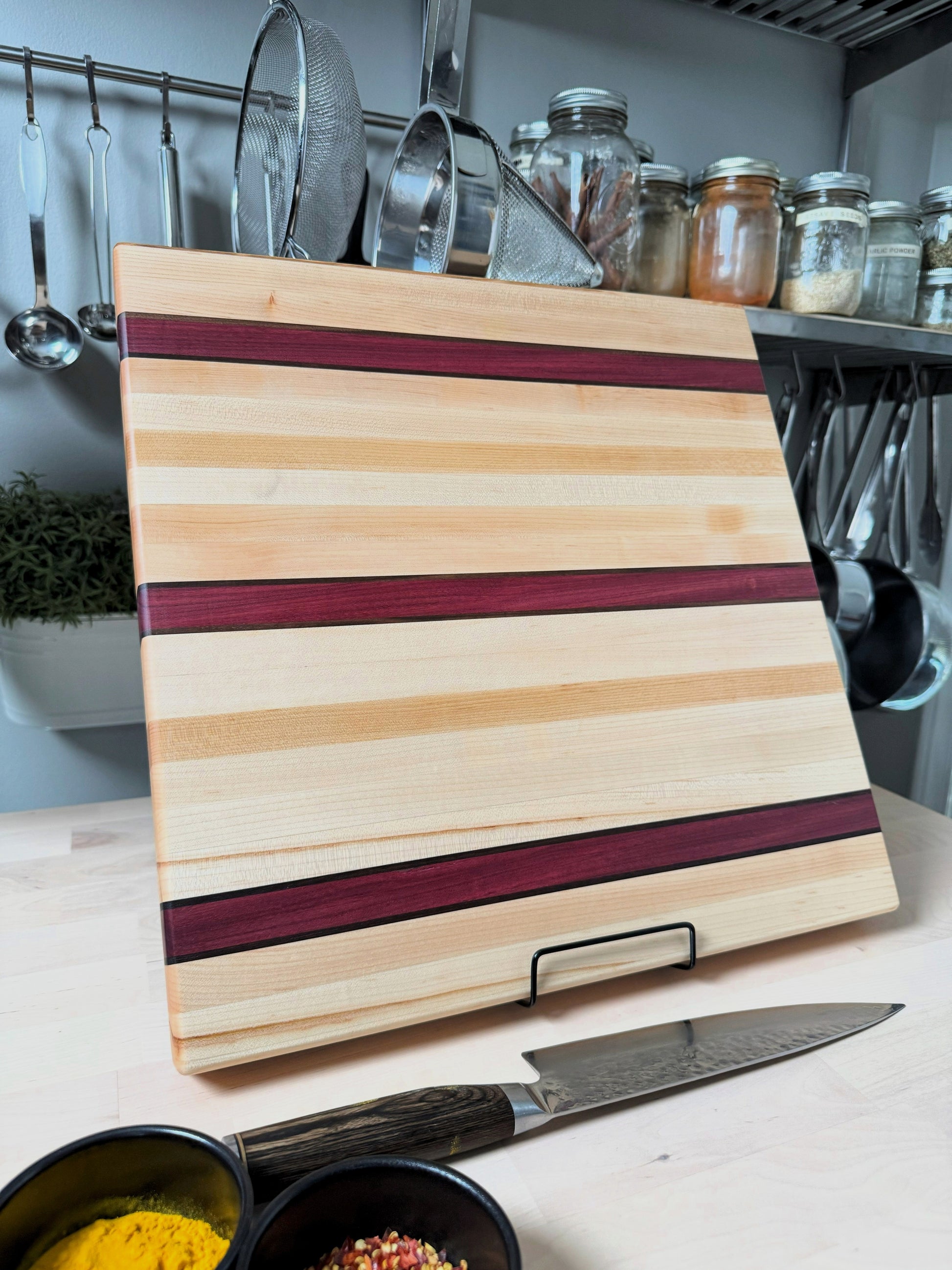 Handcrafted maple edge grain cutting board with bold Purpleheart stripes and walnut accent lines, displayed standing.