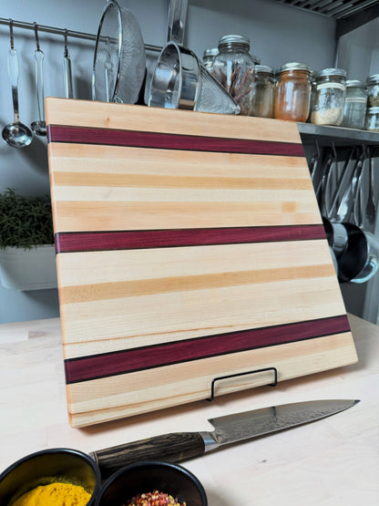Handcrafted maple edge grain cutting board with bold Purpleheart stripes and walnut accent lines, displayed standing.