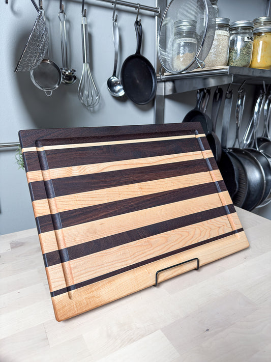 A striped wooden cutting board with alternating light and dark wood bands, resting on a kitchen counter with hanging pots and pans in the background.