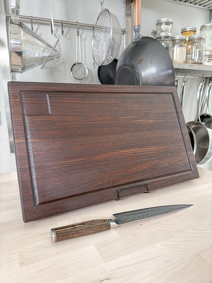 Handcrafted walnut BBQ cutting board with integrated corner drip tray, displayed standing beside a chef's knife.