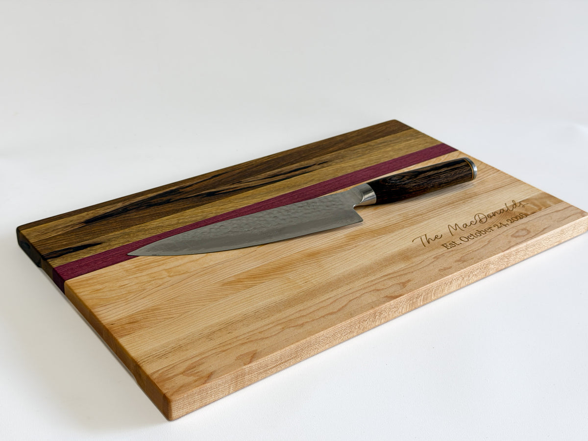 Angled view of a maple, purpleheart and black limba charcuterie board with a chef’s knife resting on its surface.