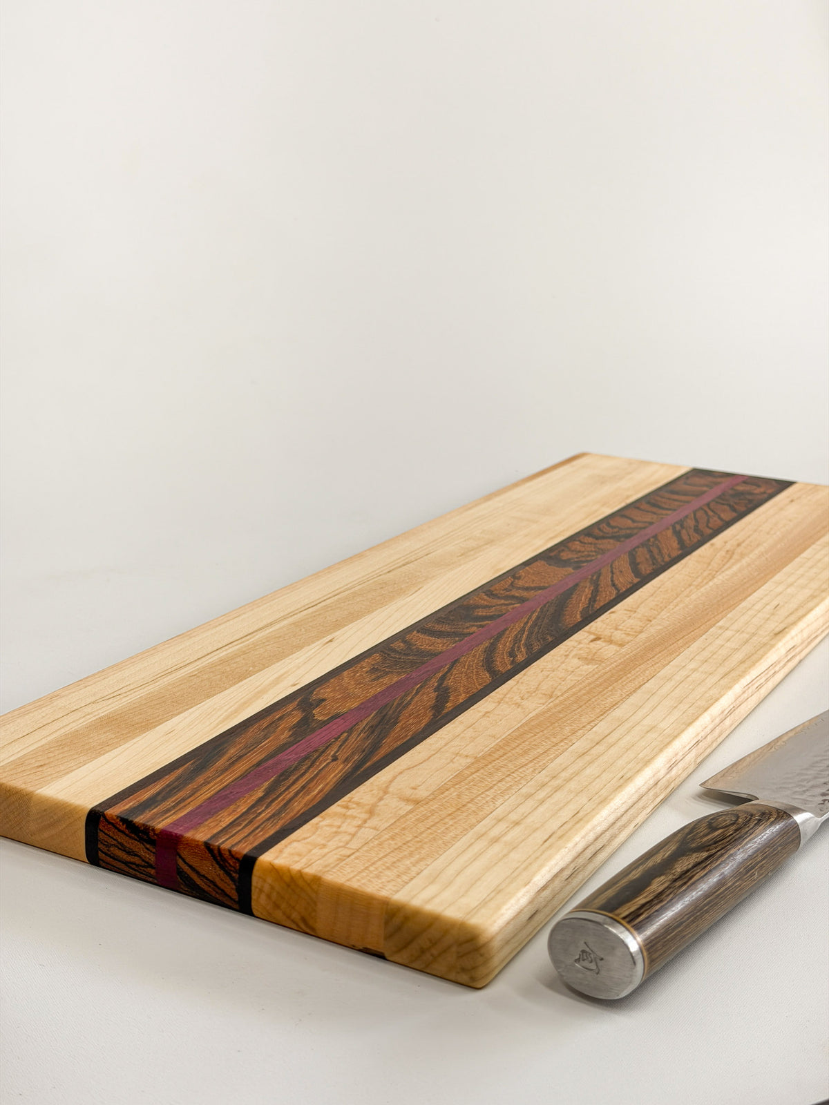 Angled close-up of the Zephyr board’s maple surface, zebrawood center, and purpleheart stripe beside a knife.