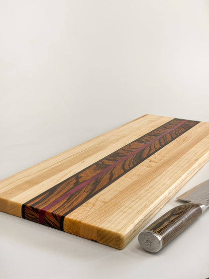 Angled close-up of the Zephyr board’s maple surface, zebrawood center, and purpleheart stripe beside a knife.