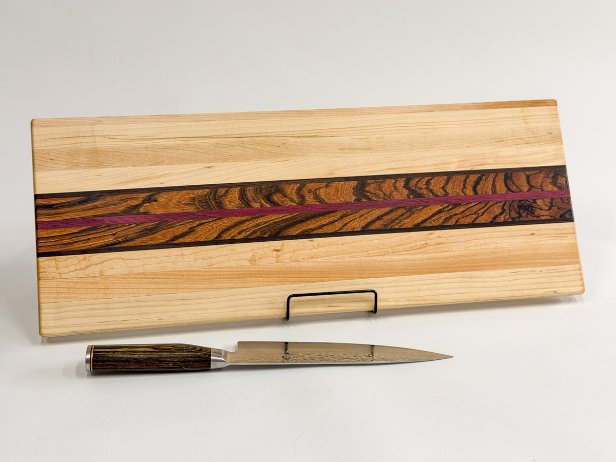 Full view of the Zephyr maple cutting board with its patterned zebrawood center, purpleheart stripe, and knife.